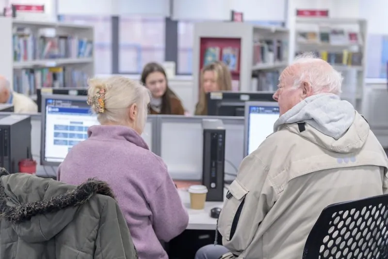 Two older adults seated at computers in foreground, one wearing a light jacket and the other in a purple fleece with a hair clip; behind them, younger people focus on screens, with bookshelves and natural light visible
