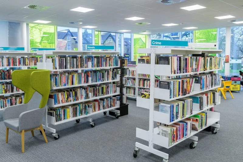 Contemporary library interior with white mobile bookshelves labelled 'Novels' and 'Crime,' large windows providing natural light, grey carpeted flooring, and a green-and-grey reading chair in the foreground