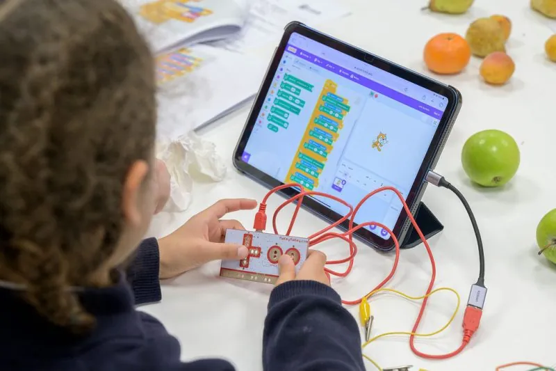 Child interacting with a Makey Makey invention kit connected to a tablet displaying Scratch programming blocks and character; wires and alligator clips link the board to various fruits on the table