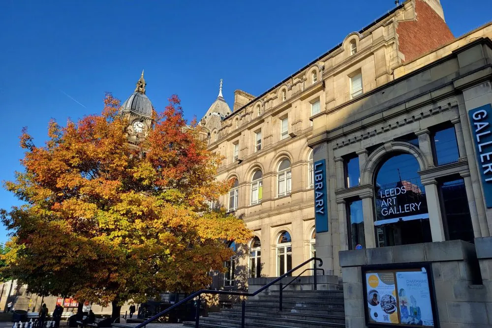 Exterior of Leeds Art Gallery and Library featuring ornate architecture and clear signage; vibrant autumn tree with multicoloured foliage contrasts against stonework, with display board and entrance steps visible