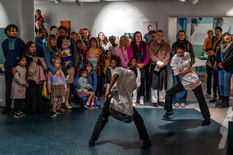Two performers in white shirts deliver an energetic dance routine in a gallery space, watched by a large crowd of adults and children gathered closely around them