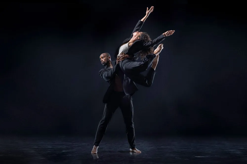 A pair of dancers execute a dynamic, mid-air pose, one holding the other close while extending their limbs gracefully against a dark, minimal background