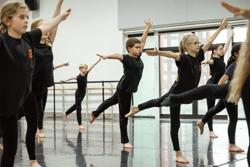 Young dancers stand barefoot in a studio, holding a controlled pose with extended arms and raised legs, reflected on the polished floor