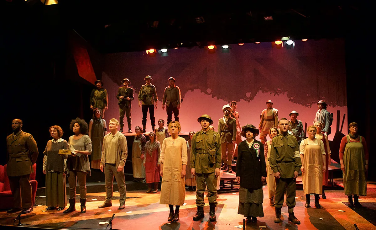 A large cast stands on stage in costume, with the front row dressed as soldiers and civilians. Behind them, more performers dressed as soldiers stand on raised platforms under dramatic lighting. The backdrop is a muted, abstract design.