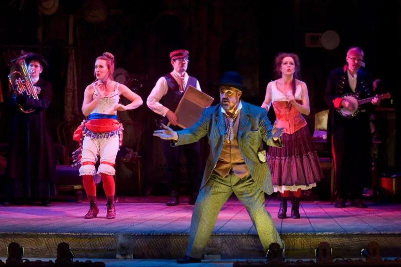 A group of six actors in colorful, vintage costumes perform on stage; one man gestures dramatically at the front, while others stand behind with musical instruments and expressive poses under theatrical lighting