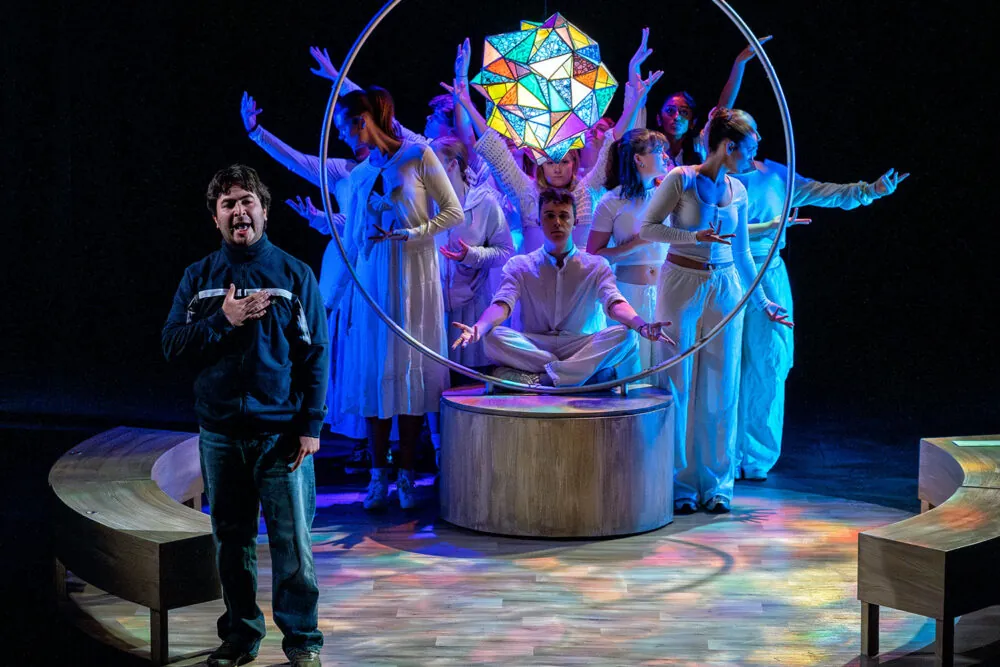 A group of actors in white costumes poses around a circular stage set with a colorful geometric light above. One person stands in front, gesturing, while others sit or stand behind in expressive poses with outstretched arms
