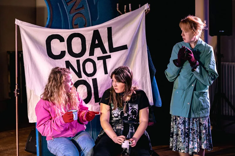 Three women talk in front of a banner reading “COAL NOT DOLE.” Two are seated, holding mugs and wearing gloves and casual clothes; the third stands, wearing a teal jacket and floral skirt, looking concerned.