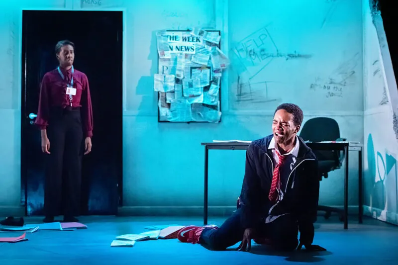 A man in a suit sits on the floor looking distressed, surrounded by scattered papers, while a woman stands in the background in front of a bulletin board labeled THE WEEK IN NEWS. The scene is lit with blue lighting
